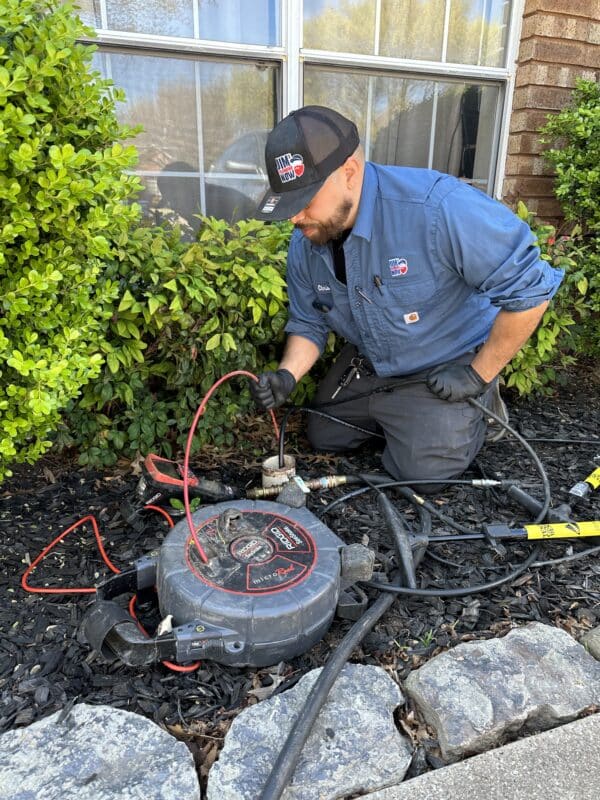 A Jim's Plumbing Now plumber in a light blue shirt kneeling down at a drain cleanout outside of a home. He is holding the line to a pinpoint camera down the drainline and looking at the camera screen.