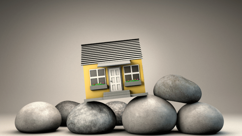 A graphic of a small yellow home sitting slightly off balance on a pile of gray stones.