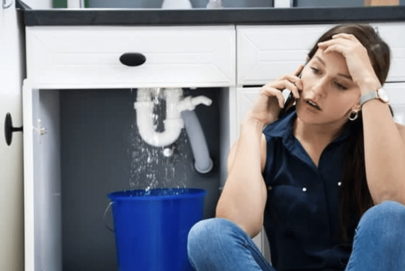 A woman sitting on the floor on the phone concerned with leaking plumbing with water dripping into a bucket behind her.