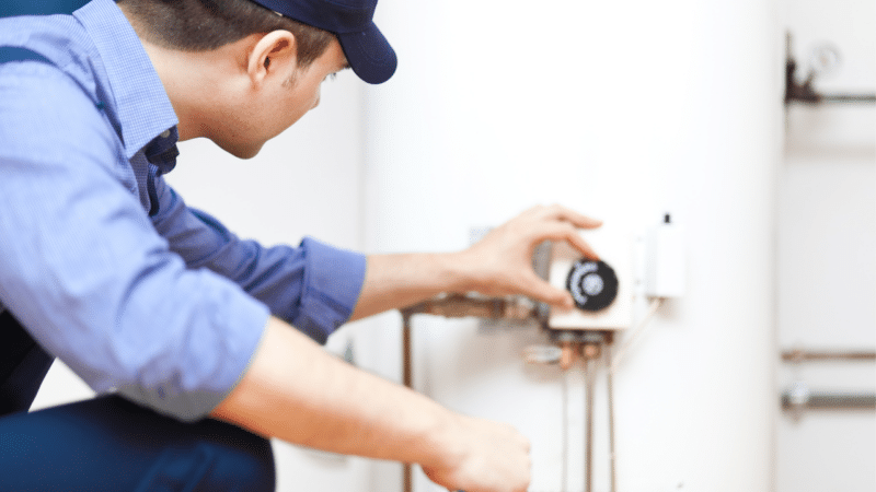 A plumber adjusting the thermostat of a traditional tank water heater.