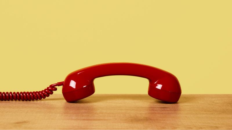 A red corded phone set down on a desk with a yellow background.